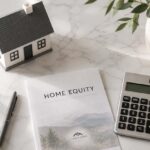 Flat lay on a clean white marble surface showing a small house model, a pen, a folded document labeled HOME EQUITY, and a calculator