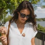 A woman in a white blouse stands outdoors on a sunny afternoon holding a rocky road ice cream cone, looking down at a large scoop that has fallen onto the front of her shirt. A waterfront railing, trees, and softly blurred scenery create a casual summer backdrop, capturing the moment an ice cream treat turns into a clothing stain