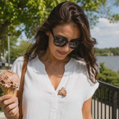 A woman in a white blouse stands outdoors on a sunny afternoon holding a rocky road ice cream cone, looking down at a large scoop that has fallen onto the front of her shirt. A waterfront railing, trees, and softly blurred scenery create a casual summer backdrop, capturing the moment an ice cream treat turns into a clothing stain