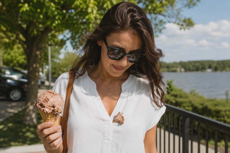 A woman in a white blouse stands outdoors on a sunny afternoon holding a rocky road ice cream cone, looking down at a large scoop that has fallen onto the front of her shirt. A waterfront railing, trees, and softly blurred scenery create a casual summer backdrop, capturing the moment an ice cream treat turns into a clothing stain