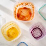 Three plastic food storage containers arranged in a loose triangle, each showing a distinct stain: one with an orange-red tomato stain on the interior walls, one with a vivid yellow turmeric stain pooled at the bottom, one with a faint purple-blue berry ring around the inside.
