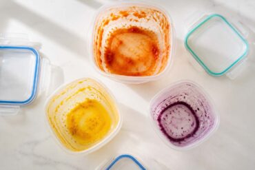 Three plastic food storage containers arranged in a loose triangle, each showing a distinct stain: one with an orange-red tomato stain on the interior walls, one with a vivid yellow turmeric stain pooled at the bottom, one with a faint purple-blue berry ring around the inside.