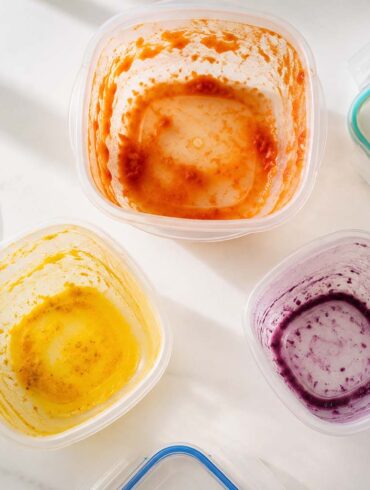Three plastic food storage containers arranged in a loose triangle, each showing a distinct stain: one with an orange-red tomato stain on the interior walls, one with a vivid yellow turmeric stain pooled at the bottom, one with a faint purple-blue berry ring around the inside.