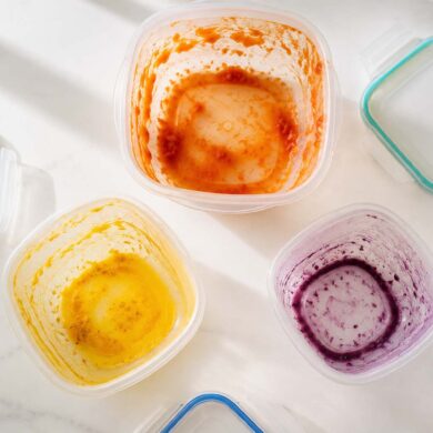 Three plastic food storage containers arranged in a loose triangle, each showing a distinct stain: one with an orange-red tomato stain on the interior walls, one with a vivid yellow turmeric stain pooled at the bottom, one with a faint purple-blue berry ring around the inside.