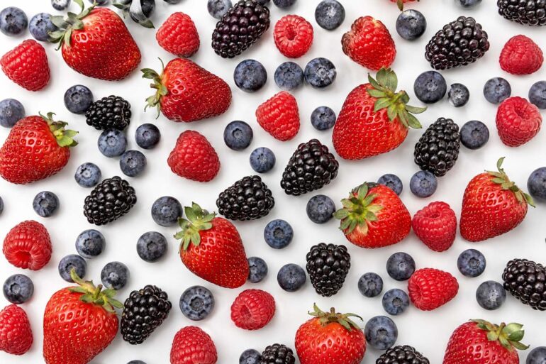 A flat lay of fresh strawberries, blueberries, raspberries, and blackberries on a white background, all unwashed and dry