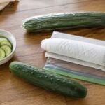  one whole unwrapped slicing cucumber, one wrapped in a paper towel beside a zip-top bag, and one English cucumber still in its plastic sleeve, with a small bowl of sliced cucumber rounds nearby