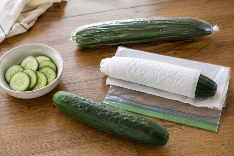 Three cucumbers on a wood surface: one whole unwrapped slicing cucumber, one wrapped in a paper towel beside a zip-top bag, and one English cucumber still in its plastic sleeve, with a small bowl of sliced cucumber rounds nearby