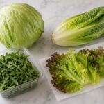Four types of lettuce on a clean marble surface: a whole head of iceberg, a romaine heart, loose leaf lettuce separated into leaves on a paper towel, and a sealed container of washed and dried arugula