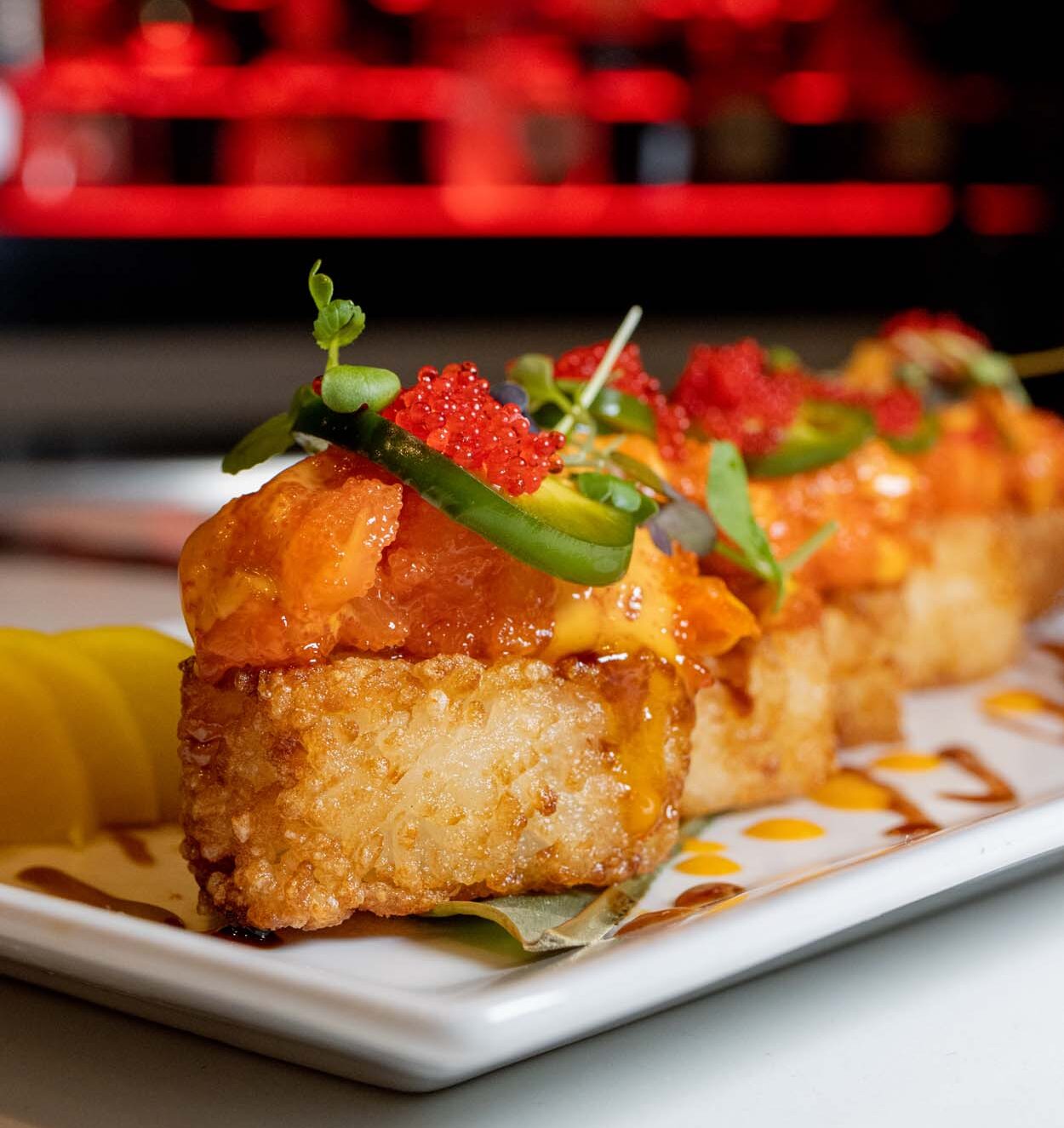 Crispy Rice Squares at LaScala's Fire topped with spicy salmon, tobiko, jalapeño, microgreens, and a drizzle of sauce, served on a white rectangular plate with the restaurant's glowing fireplace in the background