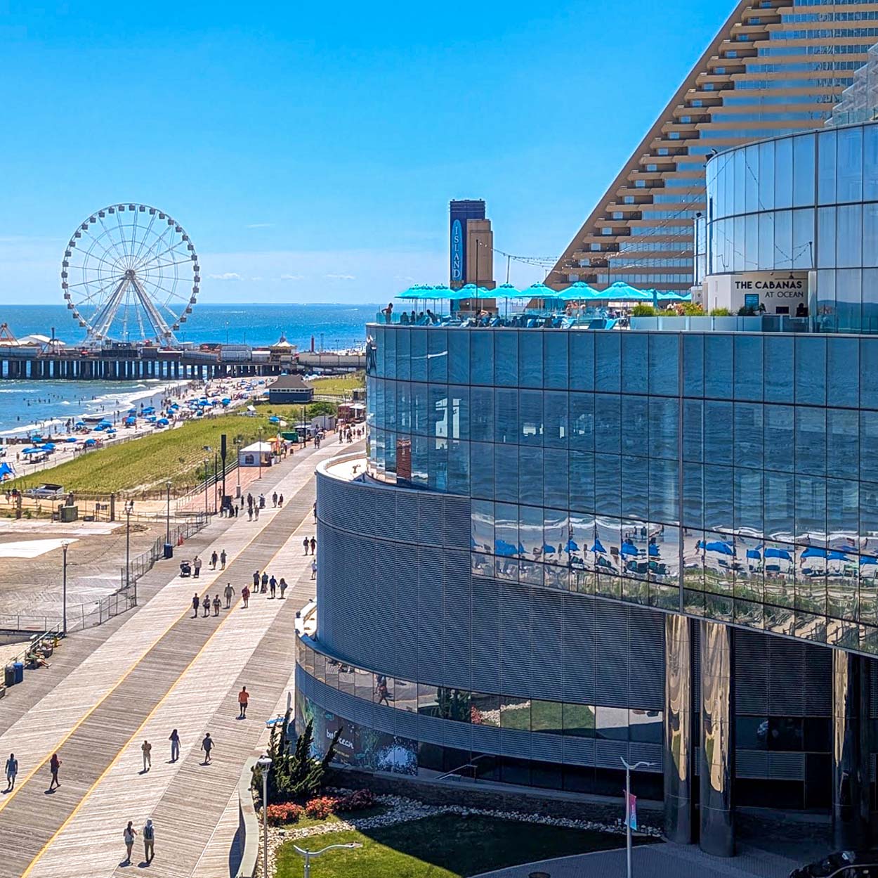 Aerial view of The Cabanas at Ocean Casino Resort on the rooftop pool deck with teal umbrellas, the Atlantic City boardwalk below, Steel Pier and the Ferris wheel in the background, and the ocean stretching to the horizon