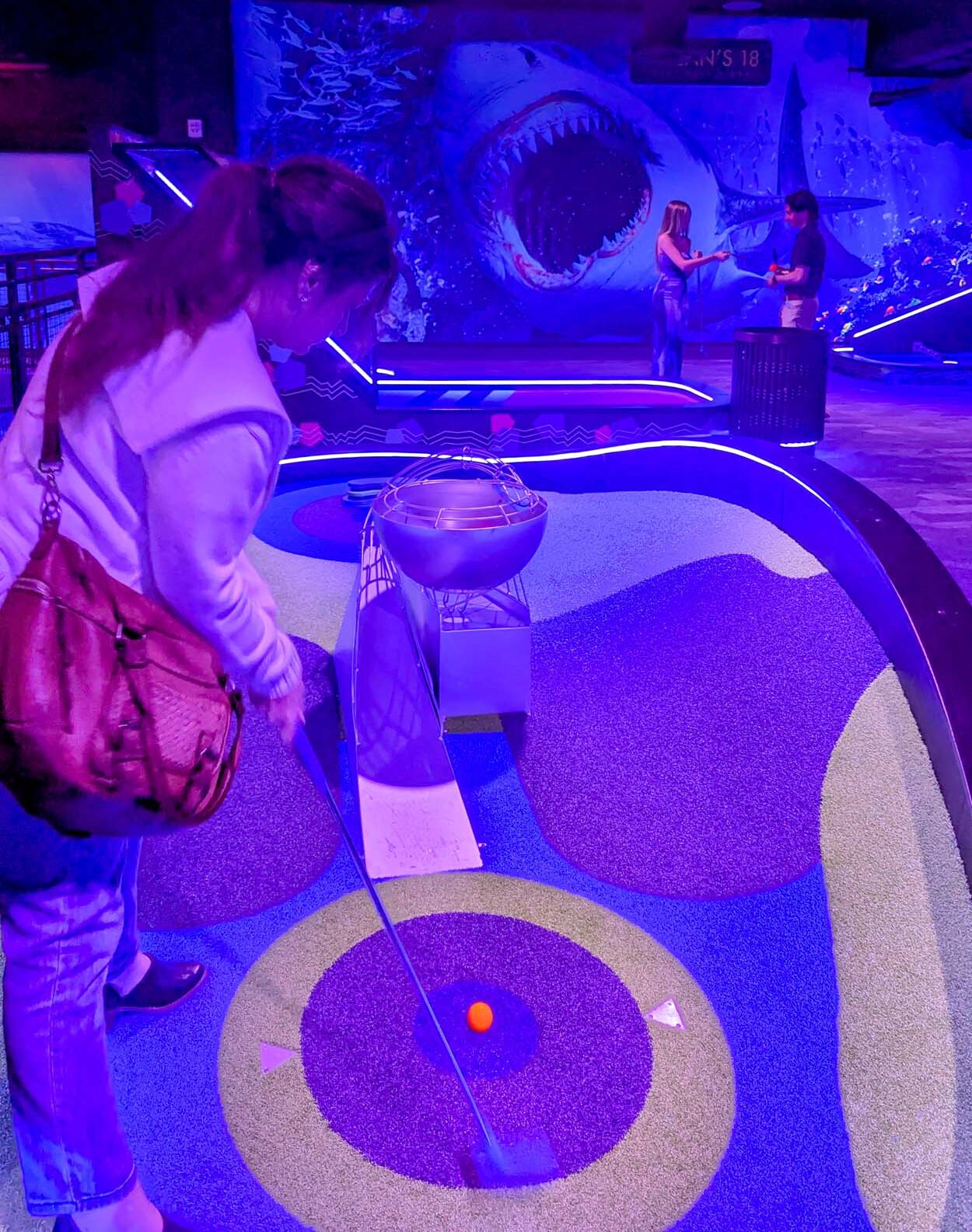 Player putting on a neon-lit hole at Ocean's 18 Mini Golf with the giant Great White Shark hologram display looming in the background