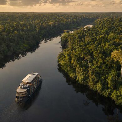 Aerial view of a small luxury expedition vessel moving slowly along a winding Amazonian river tributary, dense green rainforest canopy on both banks reflecting in the dark water, soft golden late afternoon light, no people visible, vast and untouched jungle stretching to the horizon