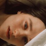 Captivating portrait of a young woman with bold eyebrows on a bed, enveloped in soft light