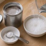 an open metal baking powder tin with its lid beside it, a small white ceramic bowl with a half teaspoon of white powder, and a glass measuring cup of steaming hot water mid-pour creating vigorous white bubbles on contact.