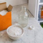 Flat lay of an open orange baking soda box beside a small white bowl of bubbling baking soda and vinegar on a white marble countertop, with a glass bottle of white vinegar and an open refrigerator showing a second baking soda box used as a deodorizer in the background.