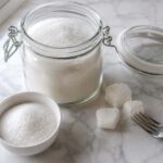 a glass airtight canister filled with white granulated sugar, lid beside it, a small white ceramic bowl with a scoop of loose sugar, and two or three visible sugar lumps next to a fork used to break them up