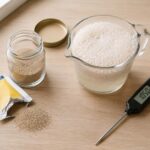 yeast packet beside a small glass jar of loose dry yeast granules, a liquid measuring cup with warm water showing a thick foam rising from a successful proofing test, and a digital kitchen thermometer resting nearby showing around 105°F
