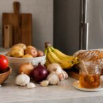 A kitchen counter with tomatoes, potatoes, onions, garlic, bananas, bread, and honey arranged outside a refrigerator