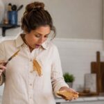 Woman in her late 30s looking down at a large peanut butter smear on the front of her light-colored shirt while holding a slice of peanut butter toast and a butter knife in a bright, modern kitchen.
