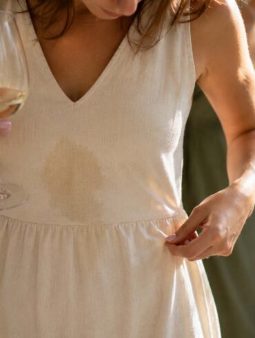 Woman in a cream linen dress at a wedding reception looking down at a fresh Sauvignon Blanc spill while holding a glass of white wine, with guests softly blurred in the background.