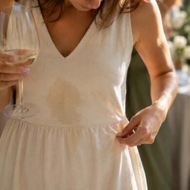Woman in a cream linen dress at a wedding reception looking down at a fresh Sauvignon Blanc spill while holding a glass of white wine, with guests softly blurred in the background.
