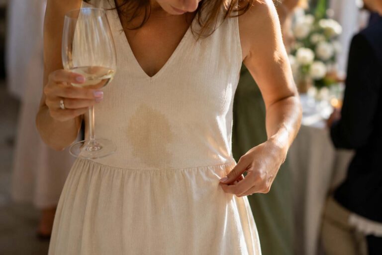 Woman in a cream linen dress at a wedding reception looking down at a fresh Sauvignon Blanc spill while holding a glass of white wine, with guests softly blurred in the background.