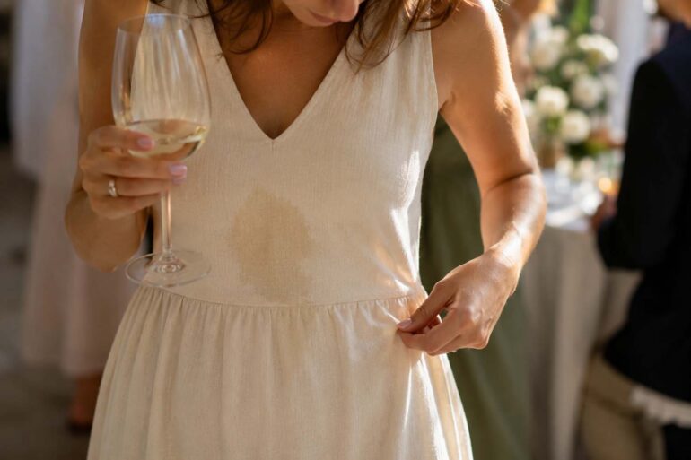Woman in a cream linen dress at a wedding reception looking down at a fresh Sauvignon Blanc spill while holding a glass of white wine, with guests softly blurred in the background.