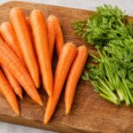 Fresh whole carrots with green tops on a wooden cutting board, green tops removed and set aside
