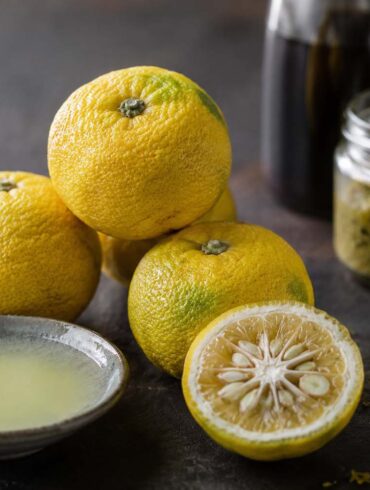 three whole yuzu fruit clustered together on a dark slate or aged wood surface — bumpy, textured yellow-green skin, slightly irregular shape, smaller than an orange. One yuzu halved to show the seedy pale yellow interior and thick pithy rind. A small ceramic dish of freshly squeezed yuzu juice beside it, pale yellow and slightly cloudy. A few pieces of freshly grated yuzu zest scattered nearby