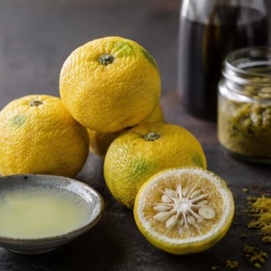 three whole yuzu fruit clustered together on a dark slate or aged wood surface — bumpy, textured yellow-green skin, slightly irregular shape, smaller than an orange. One yuzu halved to show the seedy pale yellow interior and thick pithy rind. A small ceramic dish of freshly squeezed yuzu juice beside it, pale yellow and slightly cloudy. A few pieces of freshly grated yuzu zest scattered nearby
