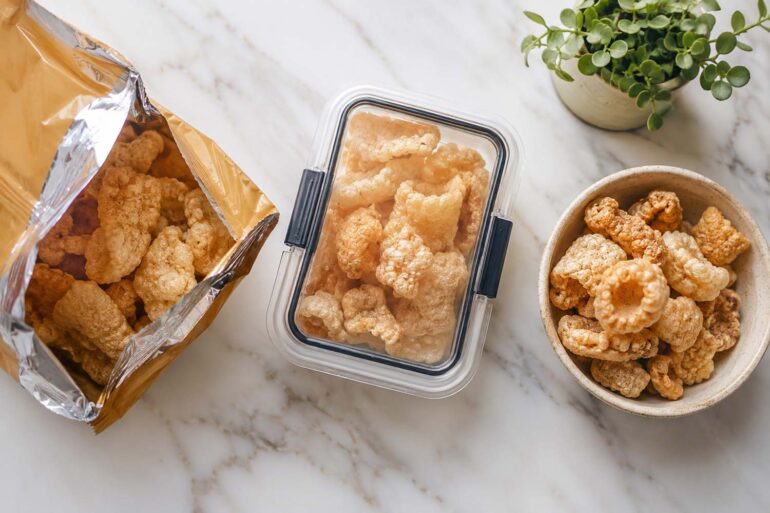 an open bag of commercial pork rinds next to an airtight storage container, alongside a small bowl of fresh chicharrones