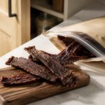 Opened resealable bag of beef jerky on a white kitchen countertop beside a small walnut cutting board with several strips of dried beef, with a softly blurred open pantry cabinet in the background illuminated by natural window light.