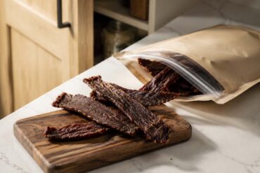 Opened resealable bag of beef jerky on a white kitchen countertop beside a small walnut cutting board with several strips of dried beef, with a softly blurred open pantry cabinet in the background illuminated by natural window light.