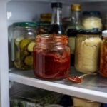 Inside an open refrigerator, an unbranded glass jar of gochujang sits among everyday condiments, with a spoonful of the thick red paste resting on the shelf nearby.