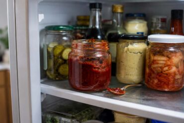 Inside an open refrigerator, an unbranded glass jar of gochujang sits among everyday condiments, with a spoonful of the thick red paste resting on the shelf nearby.