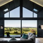 Modern living room with floor-to-ceiling A-frame windows overlooking a sunlit countryside landscape, featuring a gray sectional sofa, light wood flooring, and expansive panoramic views of rolling green fields and distant hills.