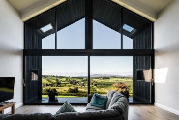 Modern living room with floor-to-ceiling A-frame windows overlooking a sunlit countryside landscape, featuring a gray sectional sofa, light wood flooring, and expansive panoramic views of rolling green fields and distant hills.