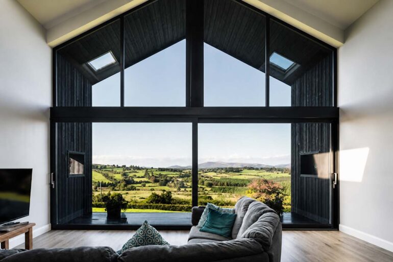 Modern living room with floor-to-ceiling A-frame windows overlooking a sunlit countryside landscape, featuring a gray sectional sofa, light wood flooring, and expansive panoramic views of rolling green fields and distant hills.