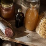 a rustic wood pantry shelf holding a honey jar, a bottle of vanilla extract, a jar of apple cider vinegar, a bag of dried pasta, and a piece of dry-cured salami