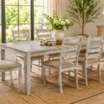 Six-piece distressed wood dining set with ladder-back chairs arranged around a rectangular dining table in a bright farmhouse-style dining room, featuring large black-framed windows, natural light, neutral decor, and a woven jute rug.