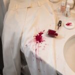Cream-colored trousers draped over a bathroom countertop with a spilled bottle of red nail polish creating a large stain on one leg beside manicure supplies and a sink.
