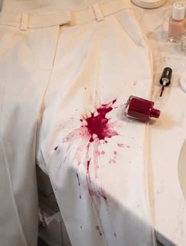 Cream-colored trousers draped over a bathroom countertop with a spilled bottle of red nail polish creating a large stain on one leg beside manicure supplies and a sink.