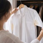 A person opening a closet and pulling out a white dress shirt on a hanger, noticing the yellow underarm stains for the first time.