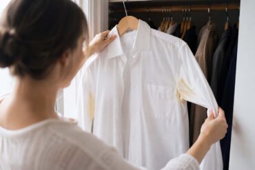 A person opening a closet and pulling out a white dress shirt on a hanger, noticing the yellow underarm stains for the first time.