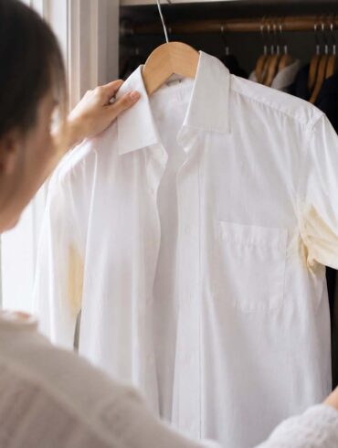 A person opening a closet and pulling out a white dress shirt on a hanger, noticing the yellow underarm stains for the first time.