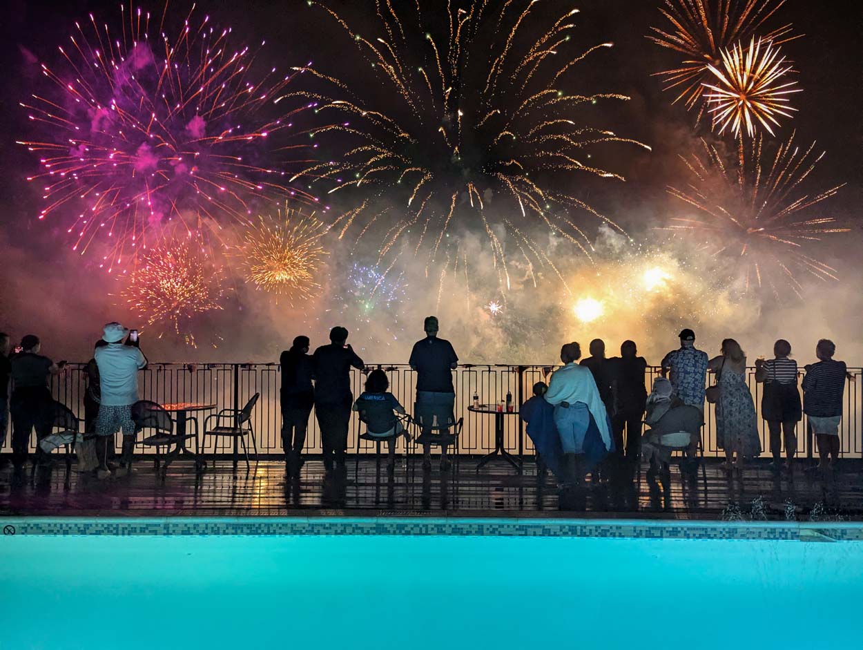 Guests watching July 4th fireworks from the rooftop pool deck at Merriweather Lakehouse Hotel in Columbia Maryland with the pool glowing teal in the foreground and fireworks reflecting on the water