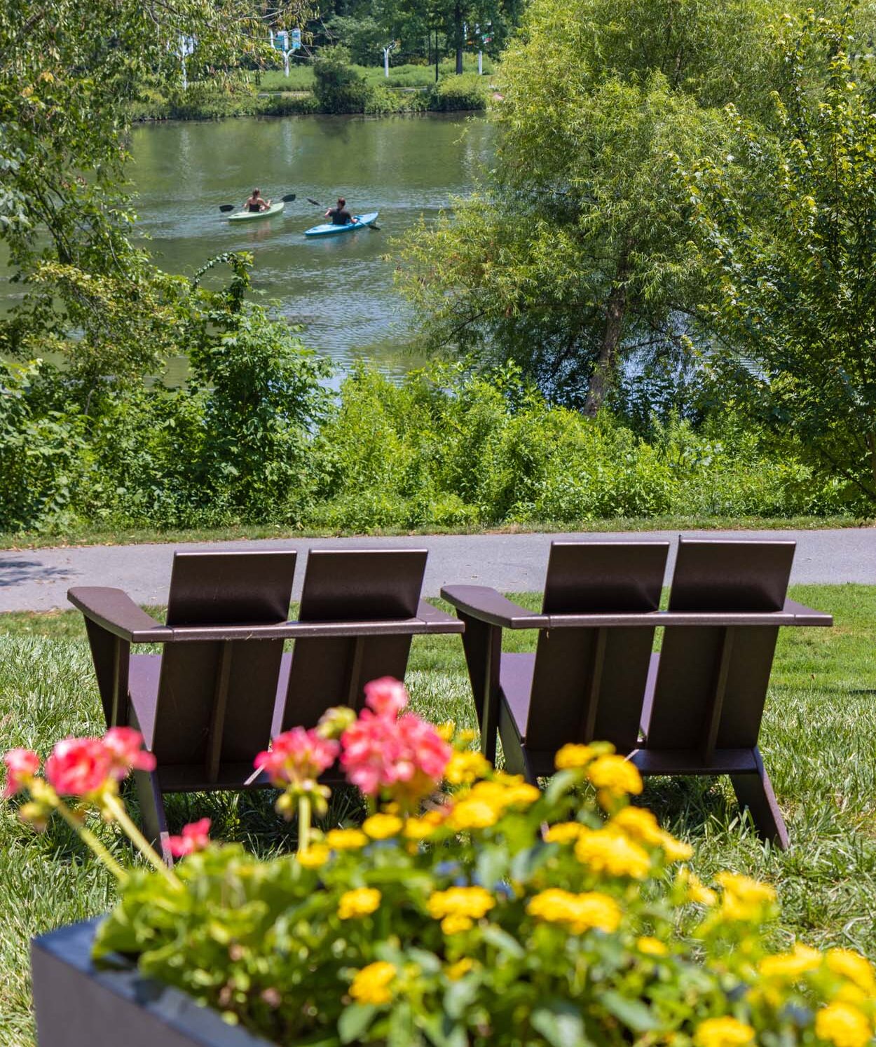 Two kayakers on Lake Kittamaqundi in summer viewed from Adirondack chairs on the grounds of Merriweather Lakehouse Hotel in Columbia Maryland with summer flowers in the foreground