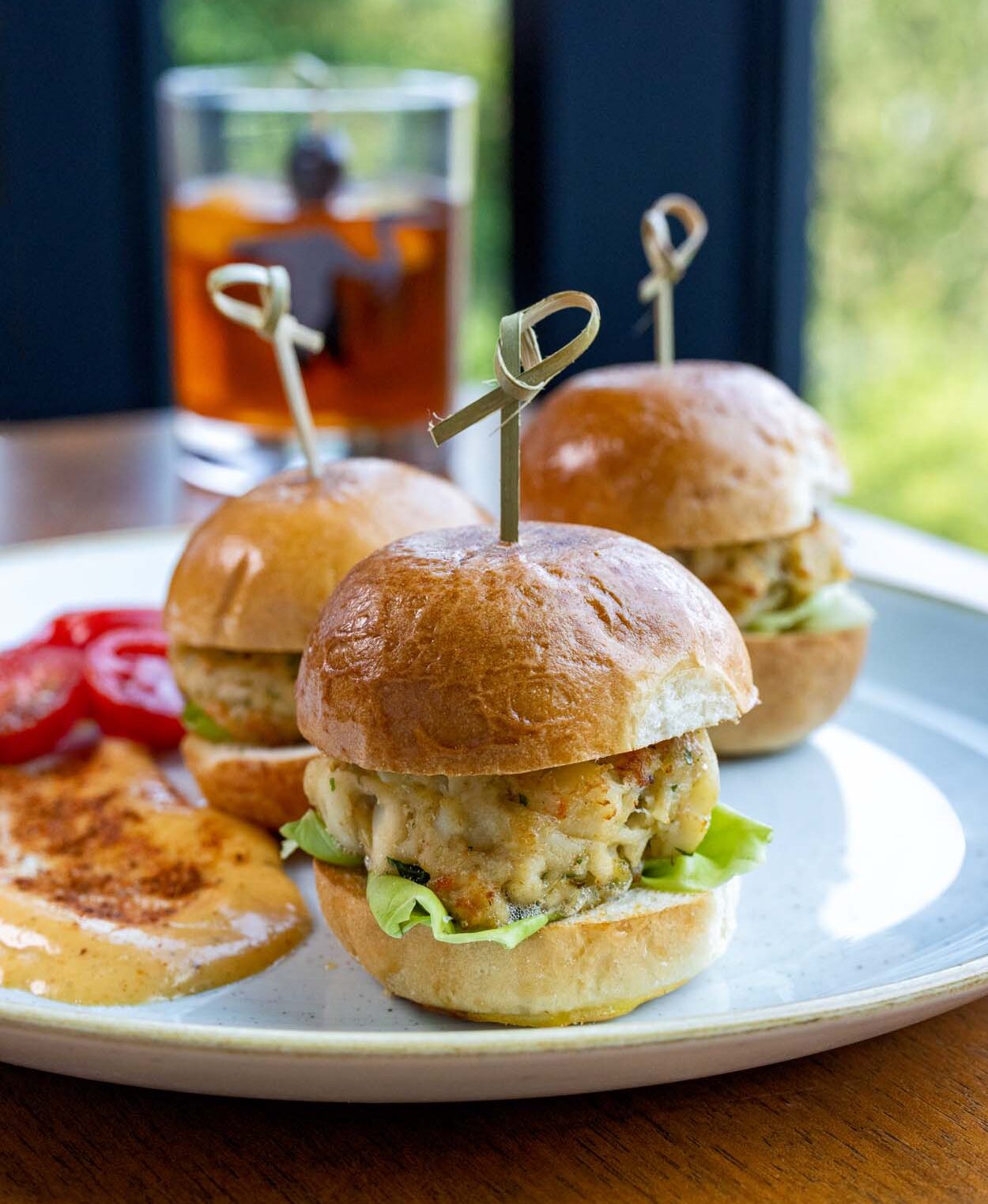 Three lump crab cake sliders on brioche buns at The Rookery Bar and Lounge at Merriweather Lakehouse Hotel with an Old Fashioned cocktail and summer green trees visible through the window