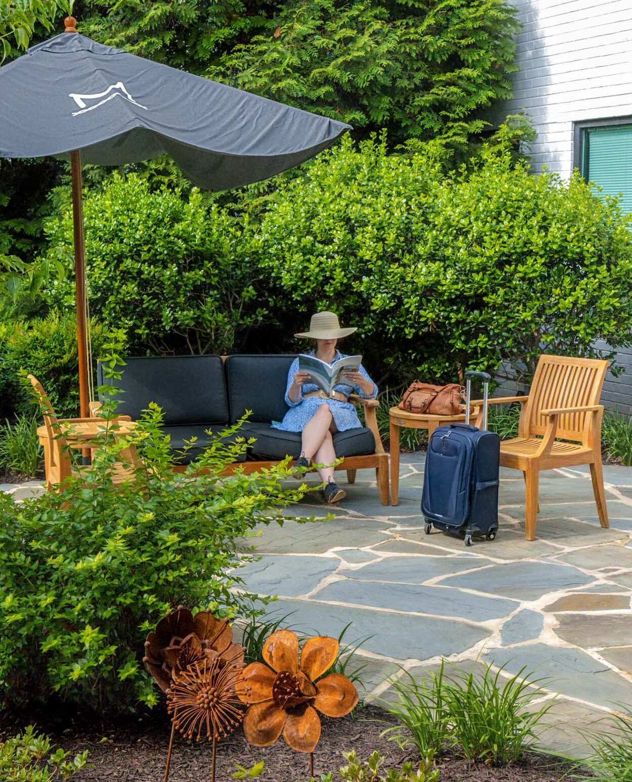 Guest in a straw hat and blue dress reading on the outdoor garden patio at Merriweather Lakehouse Hotel in Columbia Maryland with luggage and lush summer greenery surrounding the seating area