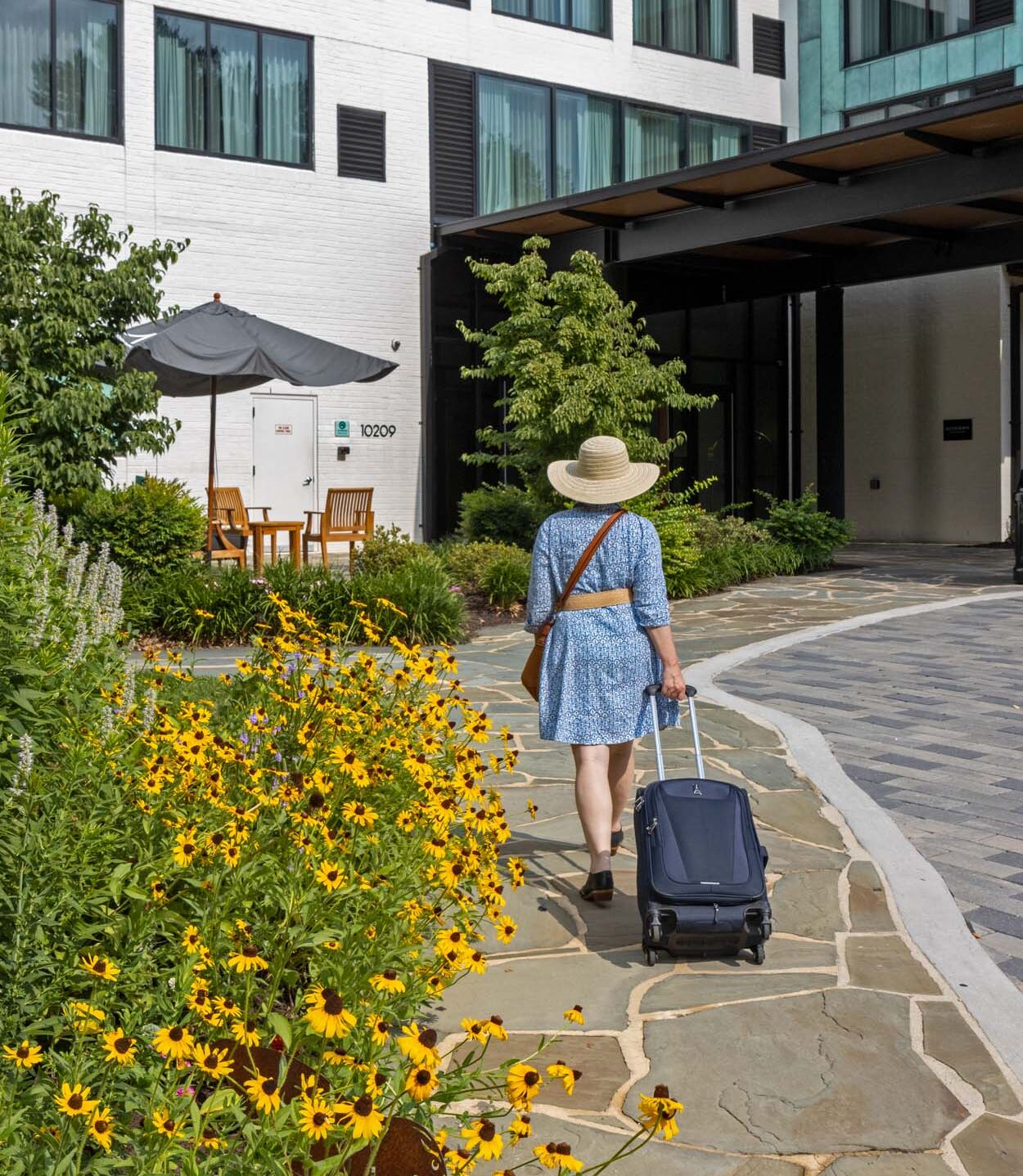 Woman in blue dress and straw hat arriving at Merriweather Lakehouse Hotel pulling luggage along a stone path lined with black eyed Susans in full summer bloom